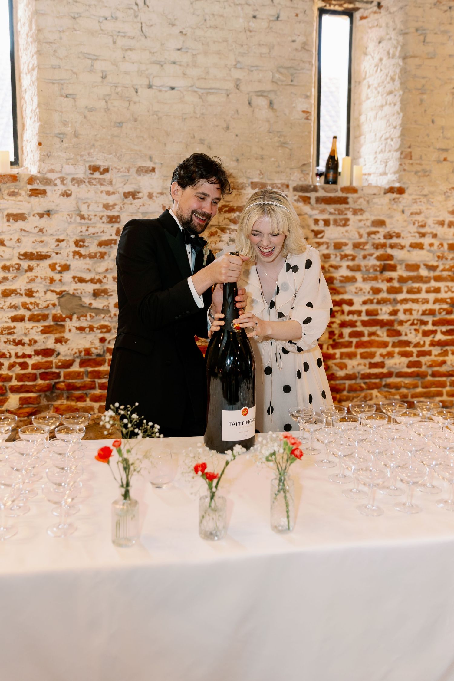 A couple celebrates joyfully against an exposed brick wall with floral arrangements in mason jars at a reception venue.