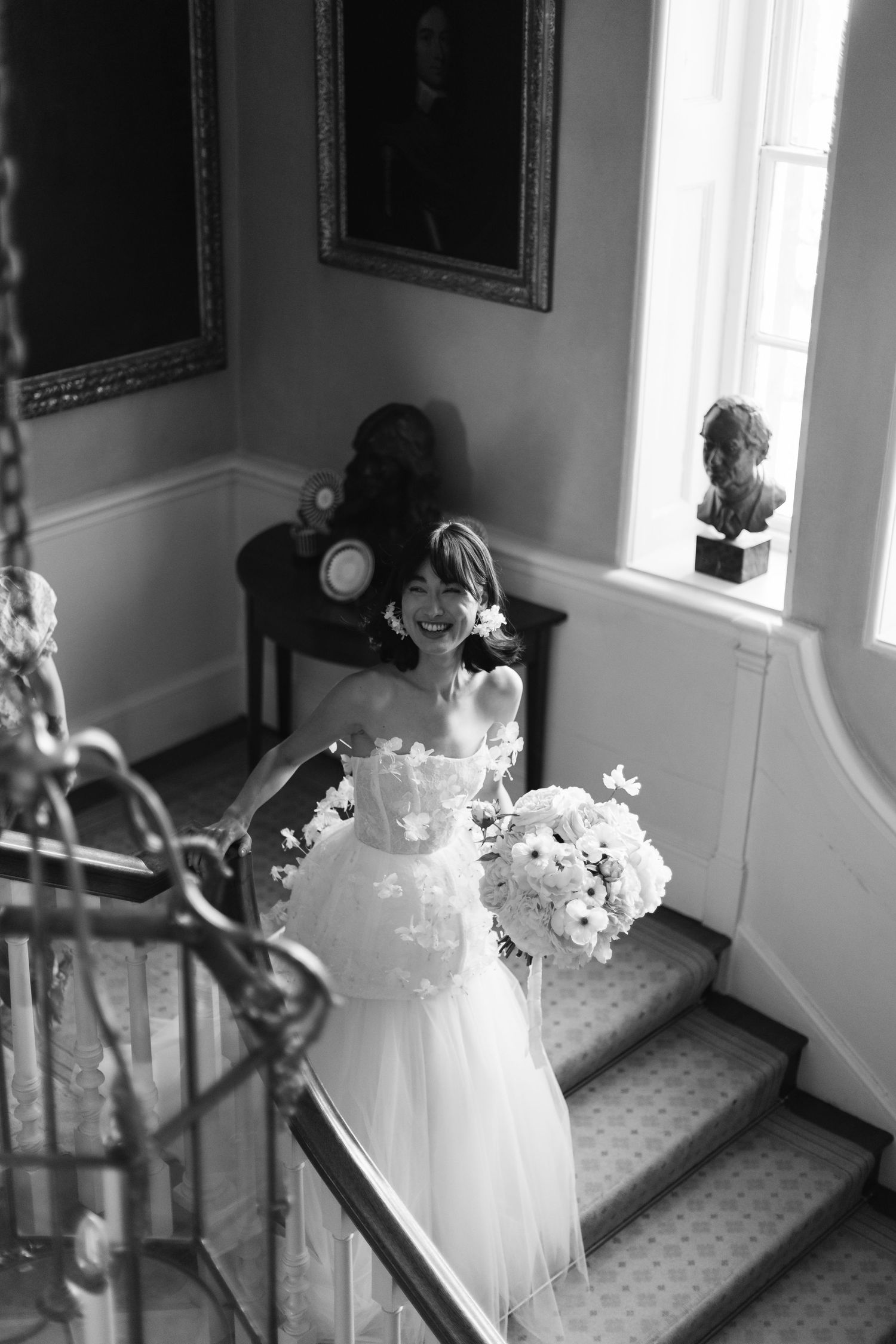 Black and white photograph of a bride in a strapless wedding gown holding a bouquet while standing on elegant stairs.