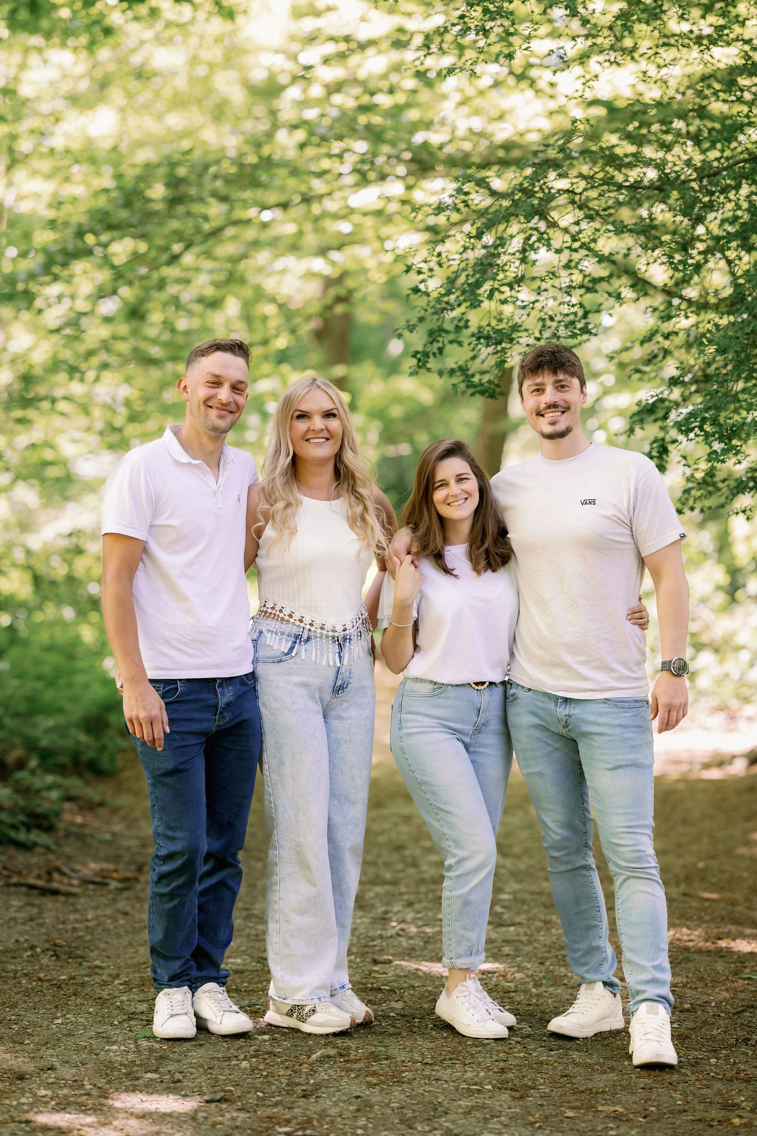 Four people in casual light-colored clothing stand together on a forest path under green leafy trees on a sunny day.