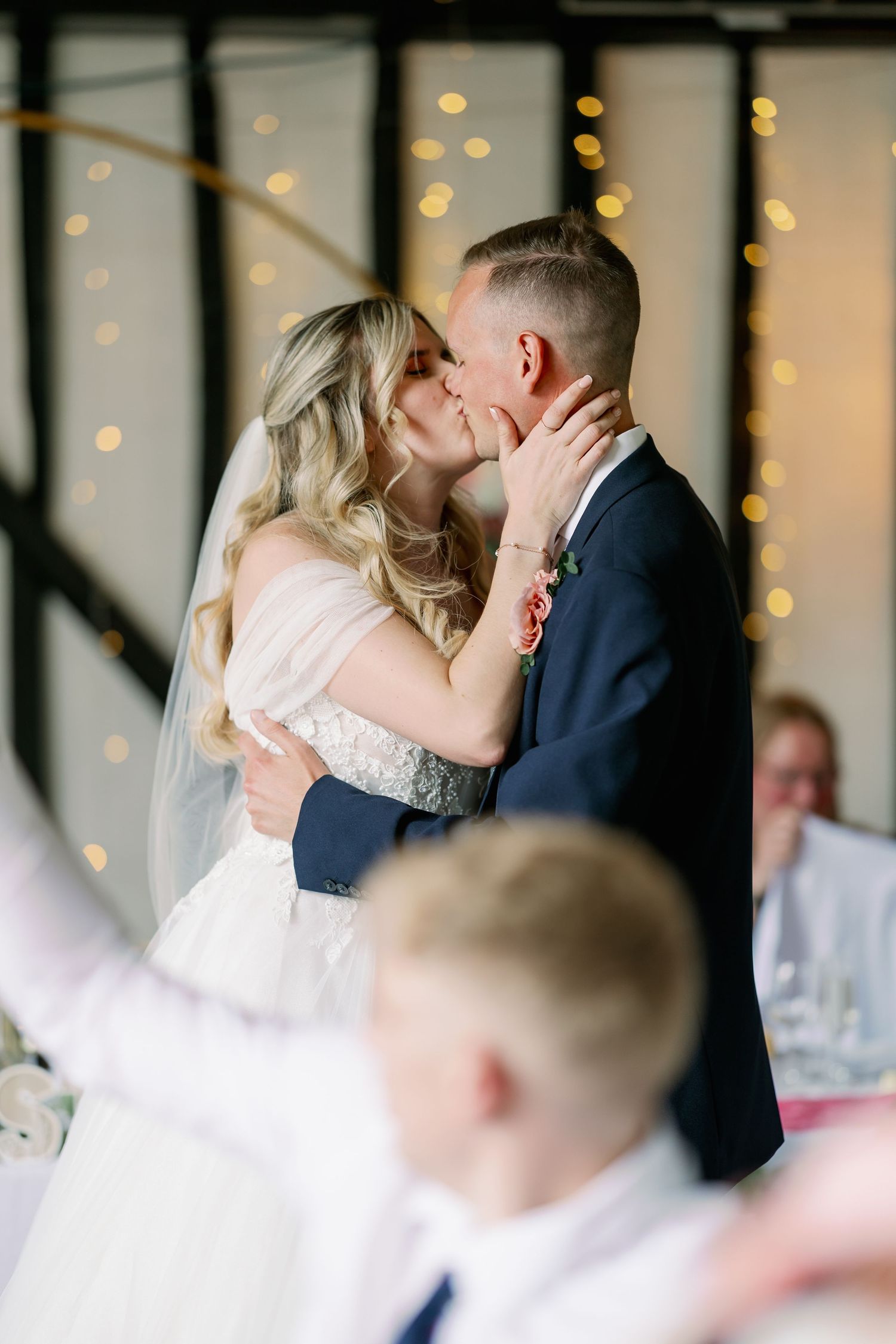 Bride and groom share a romantic kiss during their wedding reception with twinkle lights glowing in the background.