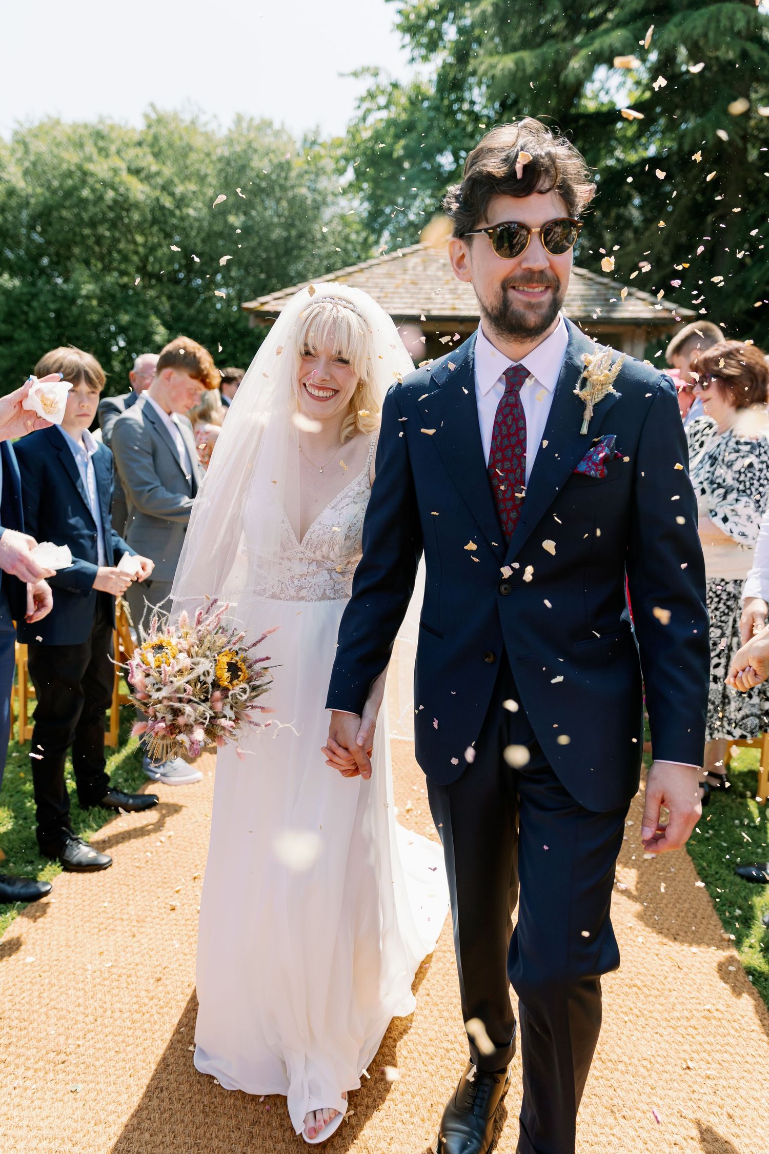 Newlywed couple walks down outdoor aisle while guests throw confetti on sunny summer day.