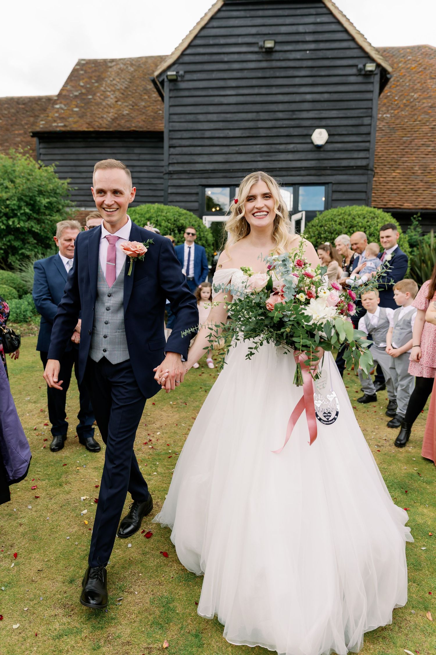 Wedding celebration on grassy lawn in front of rustic barn building with guests looking on and beautiful floral bouquet.