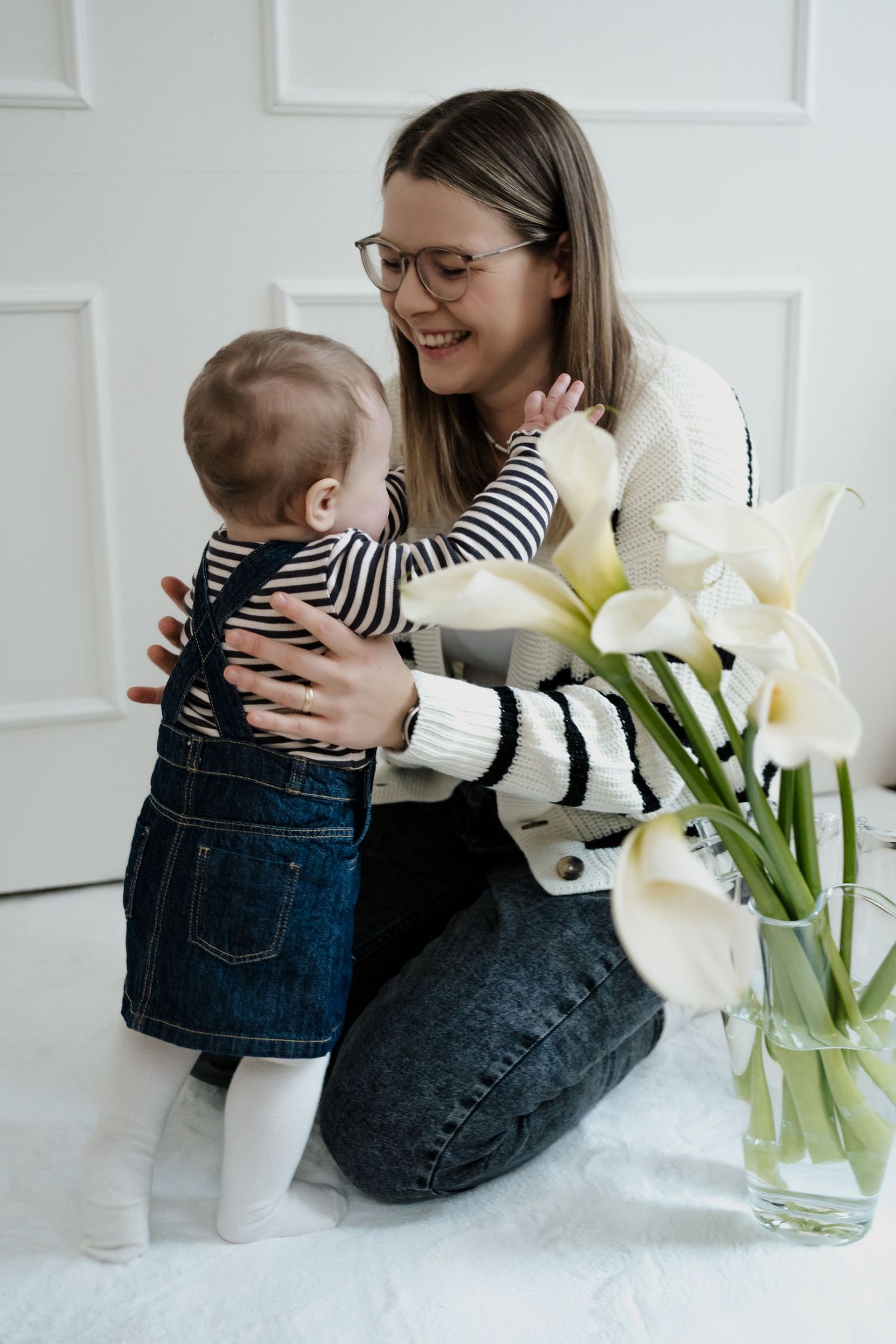 A heartwarming moment with baby in denim overalls next to white lilies in a vase on light background.