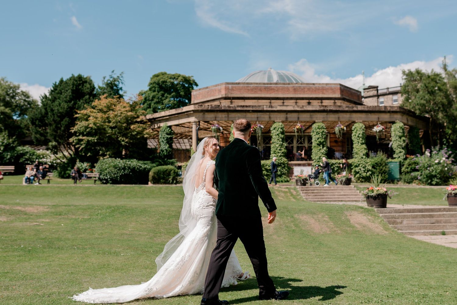 A couple walks on the green lawn in front of a historic pavilion building on a sunny summer afternoon.