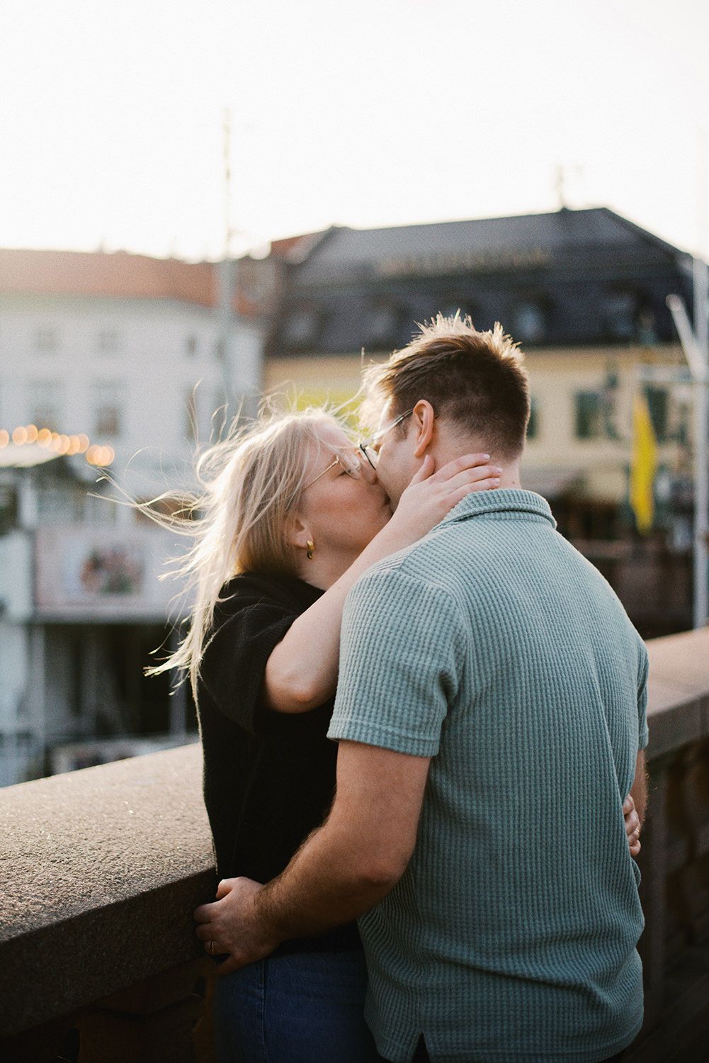 A couple shares a romantic kiss at sunset while standing on an outdoor terrace overlooking buildings.