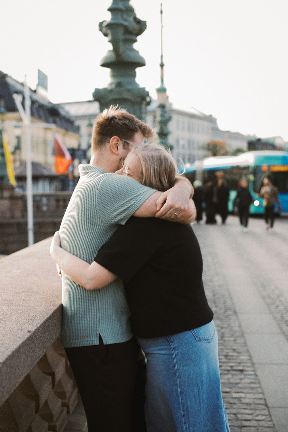 A couple sharing an intimate embrace on a city street with ornate lamp posts and buildings in the background.