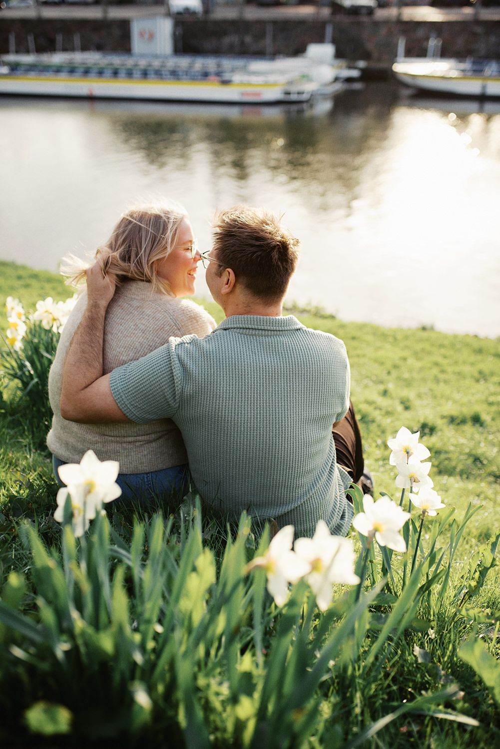 A couple sits by the riverside on a grassy bank surrounded by blooming white daffodils as a boat floats in the background.