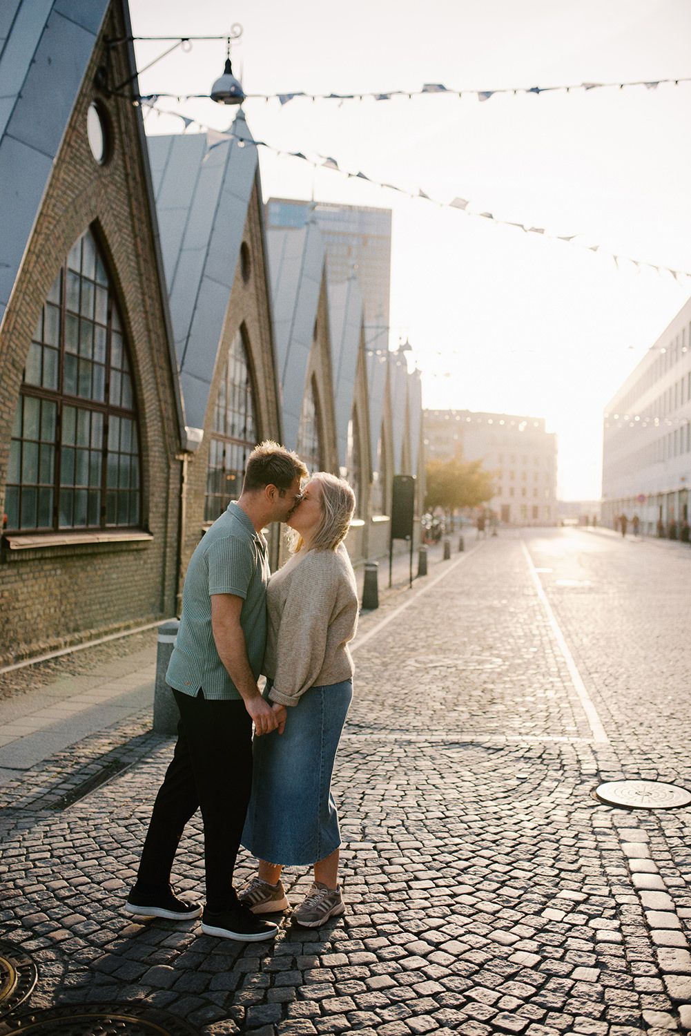 A couple shares a romantic moment on a cobblestone street at sunset with arched industrial buildings in the background.