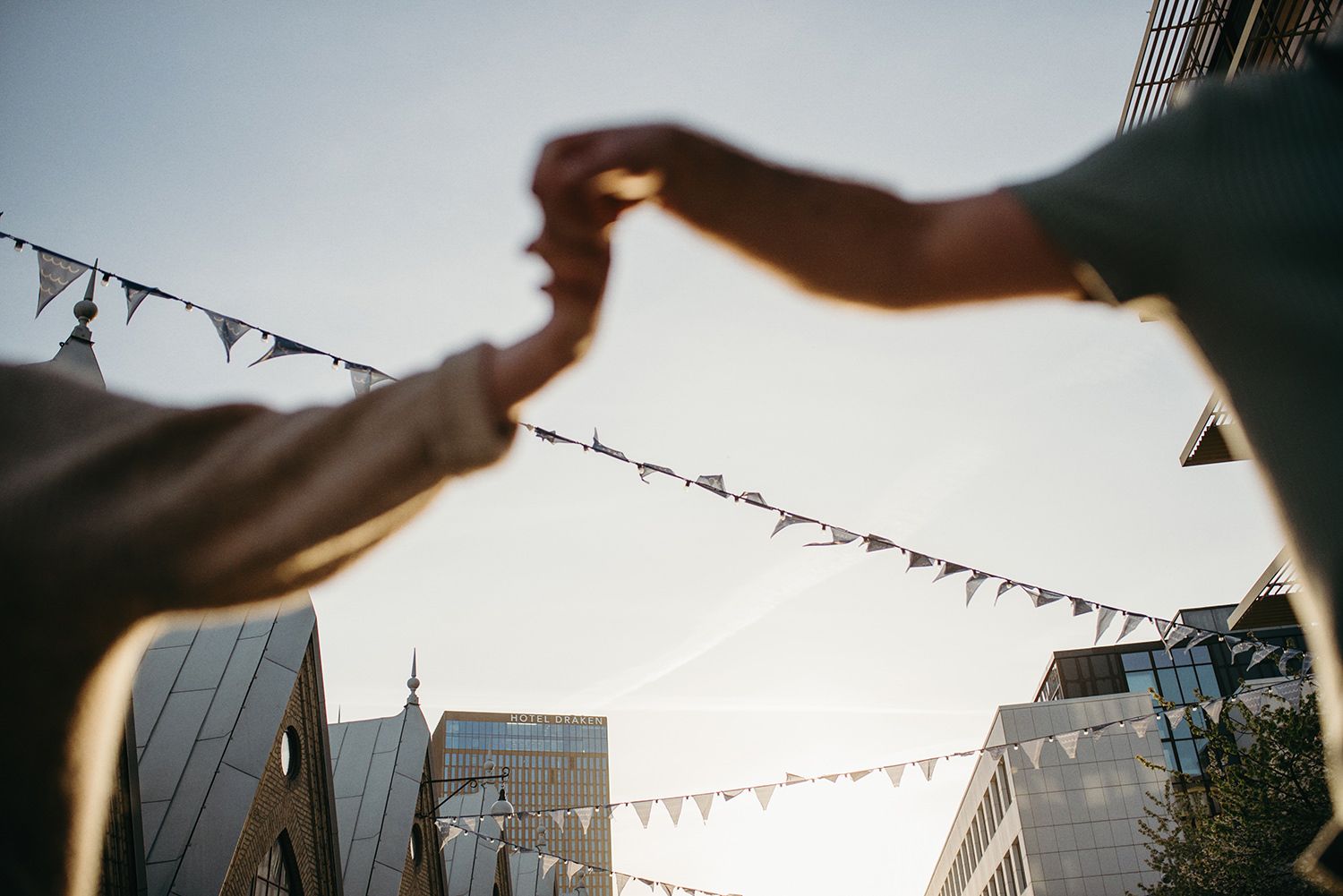 Silhouetted hands reach out beneath string lights at dusk with urban buildings in the background.