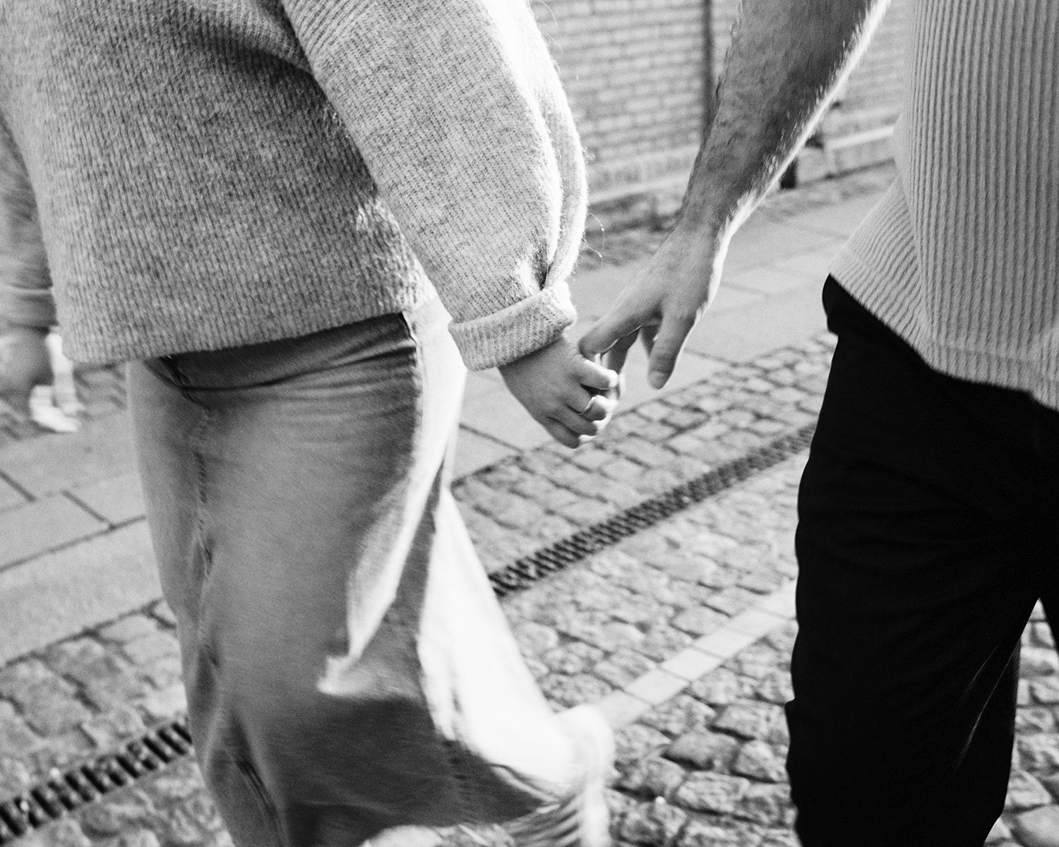 Two people hold hands while walking on a cobblestone sidewalk in black and white.