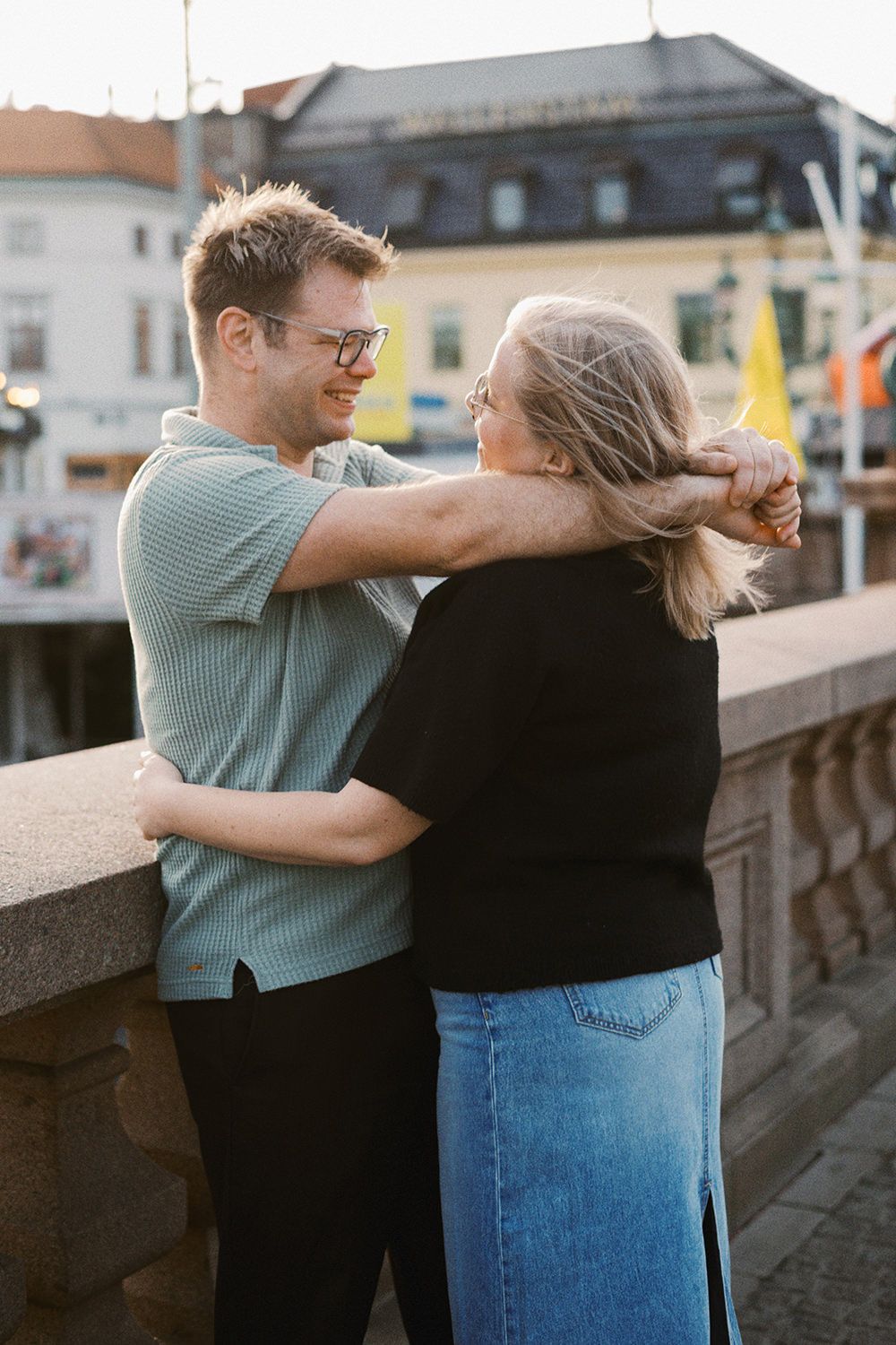 An affectionate couple shares a warm embrace while standing on a sidewalk along a waterfront during golden hour.