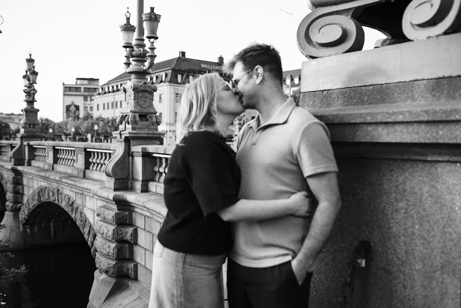 A romantic black and white photo of a couple embracing near an ornate stone bridge in a European city setting.