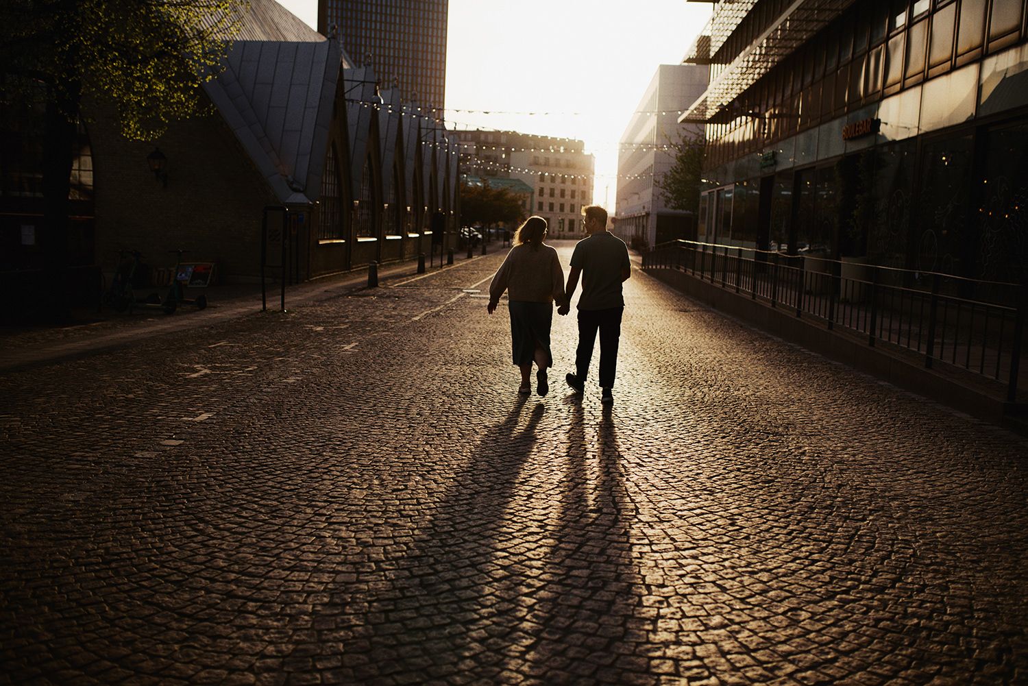 Two silhouetted figures walk hand in hand down a cobblestone street at sunset casting long shadows.