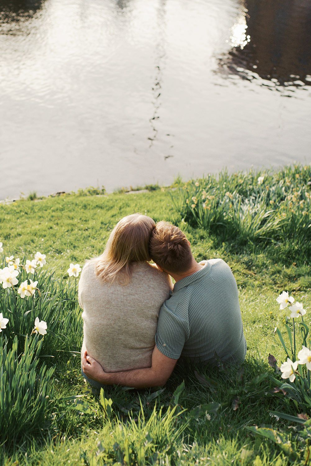 A couple sits together on a grassy bank overlooking water, surrounded by blooming white daffodils in springtime.