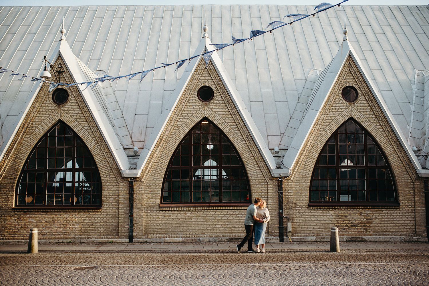 Two people stand in front of a brick church building with three pointed arches and small round windows at its peak.