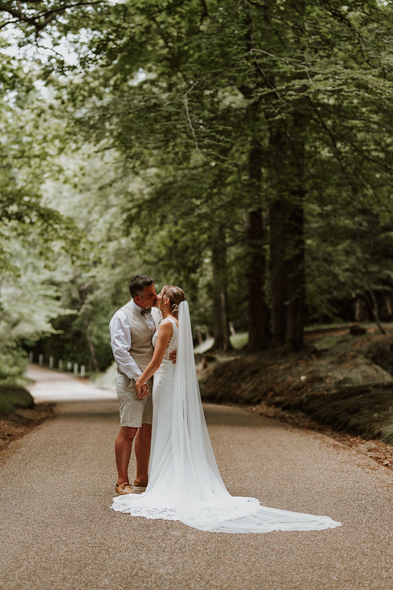 A couple shares an intimate moment on a tree-lined path with a long flowing white veil trailing behind them.