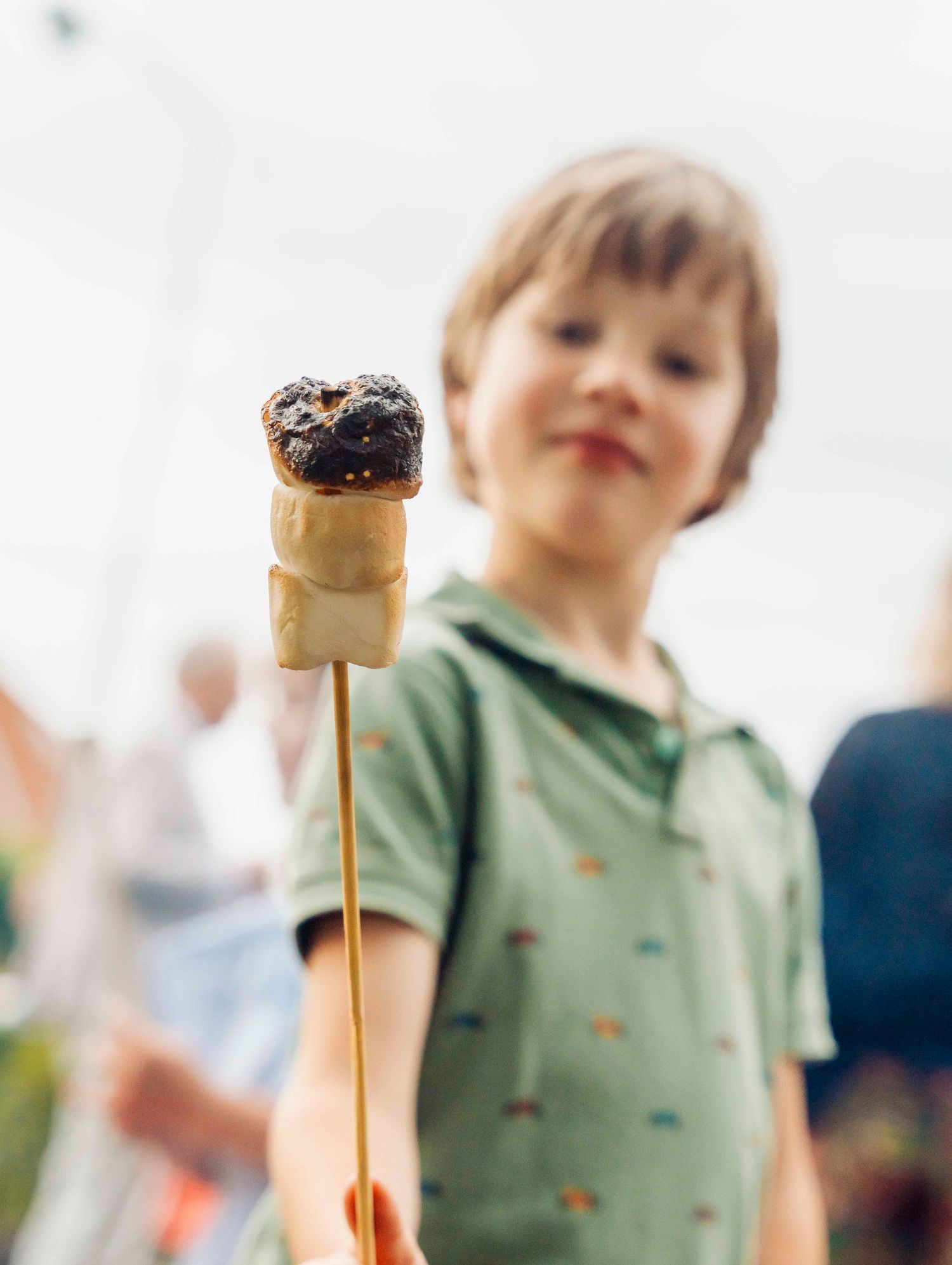 Child proudly displaying roasted marshmallow on wooden skewer against cloudy sky.