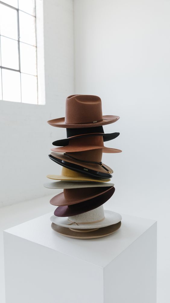 Stack of vintage fedora and cowboy hats balanced on white pedestal against bright wall.
