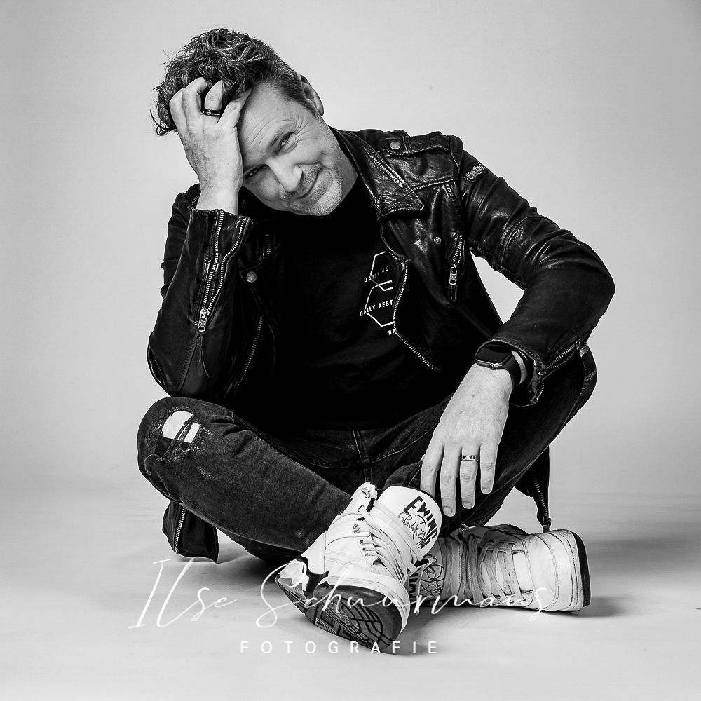 Artistic black and white portrait showing relaxed pose in leather jacket and casual attire.