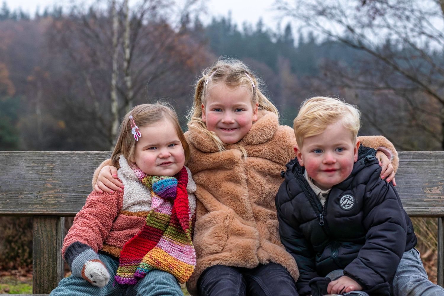 Three children sitting together on a wooden bench wearing winter coats on a cloudy day.