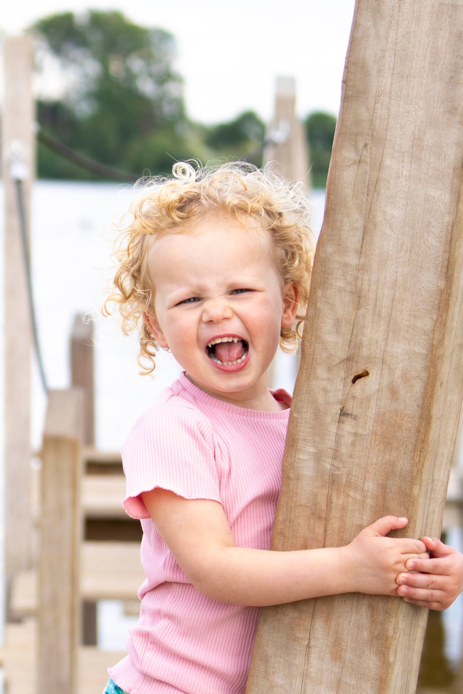 Laughing toddler in pink shirt holding onto wooden post outdoors.