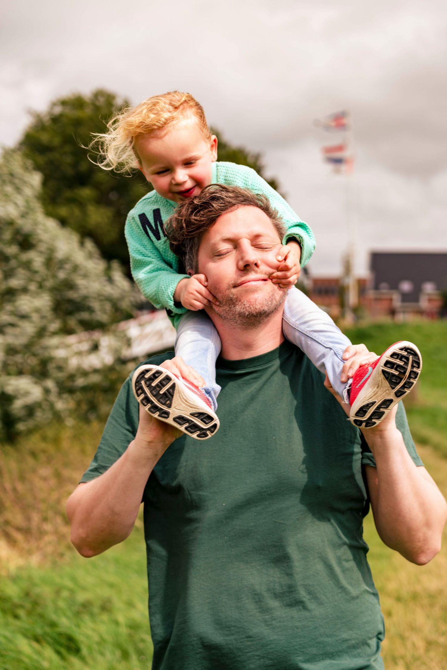 A playful parent-child shoulder ride moment in a green field with trees and cloudy skies.