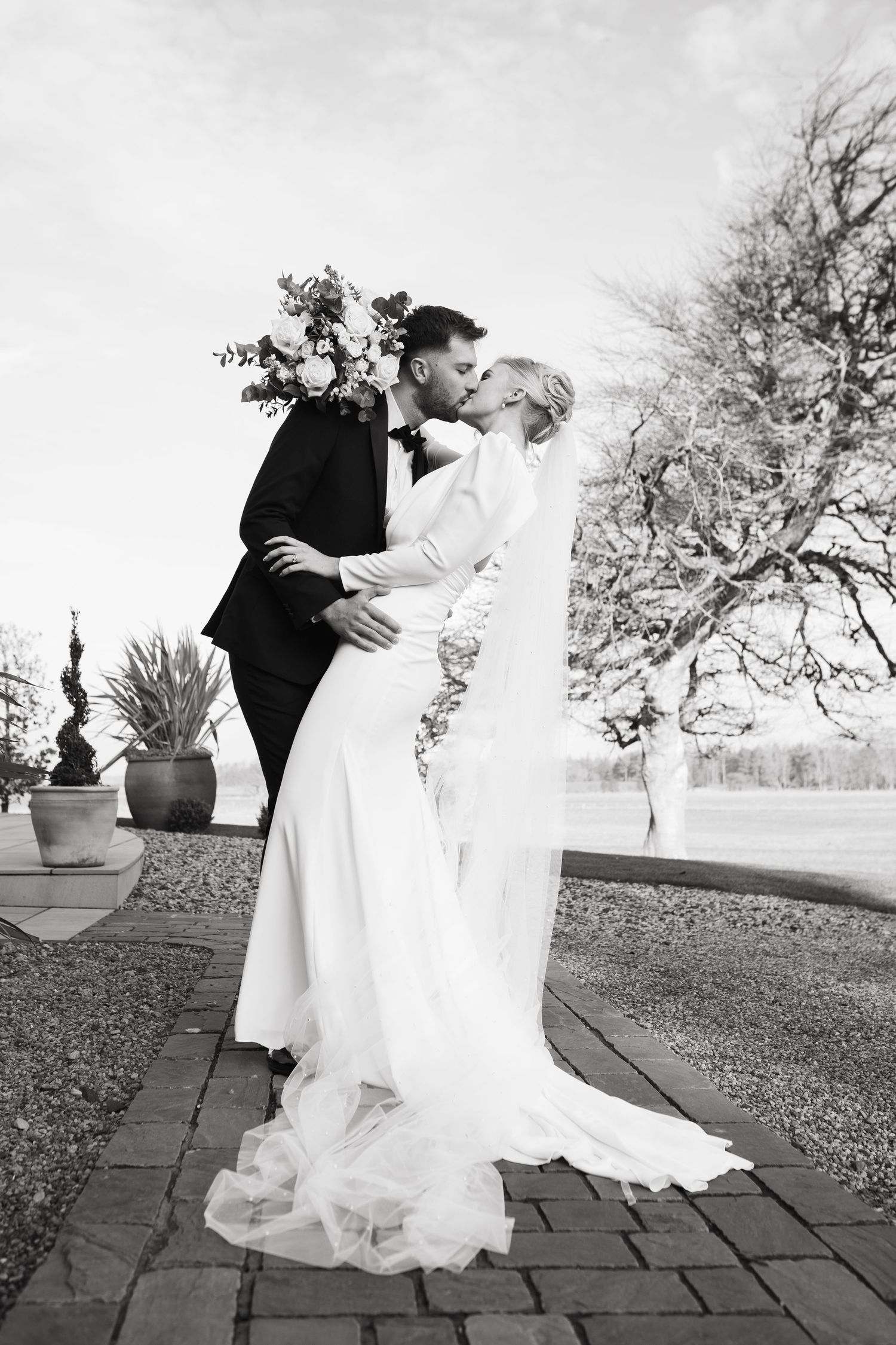 A romantic black and white wedding photo of a couple embracing outdoors on a brick patio with bare trees in background.
