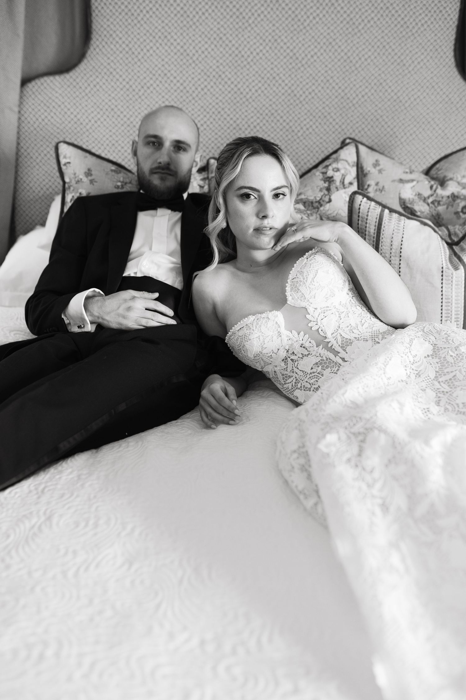 A newlywed couple relaxes together on an elegant couch in formal wedding attire, captured in black and white.