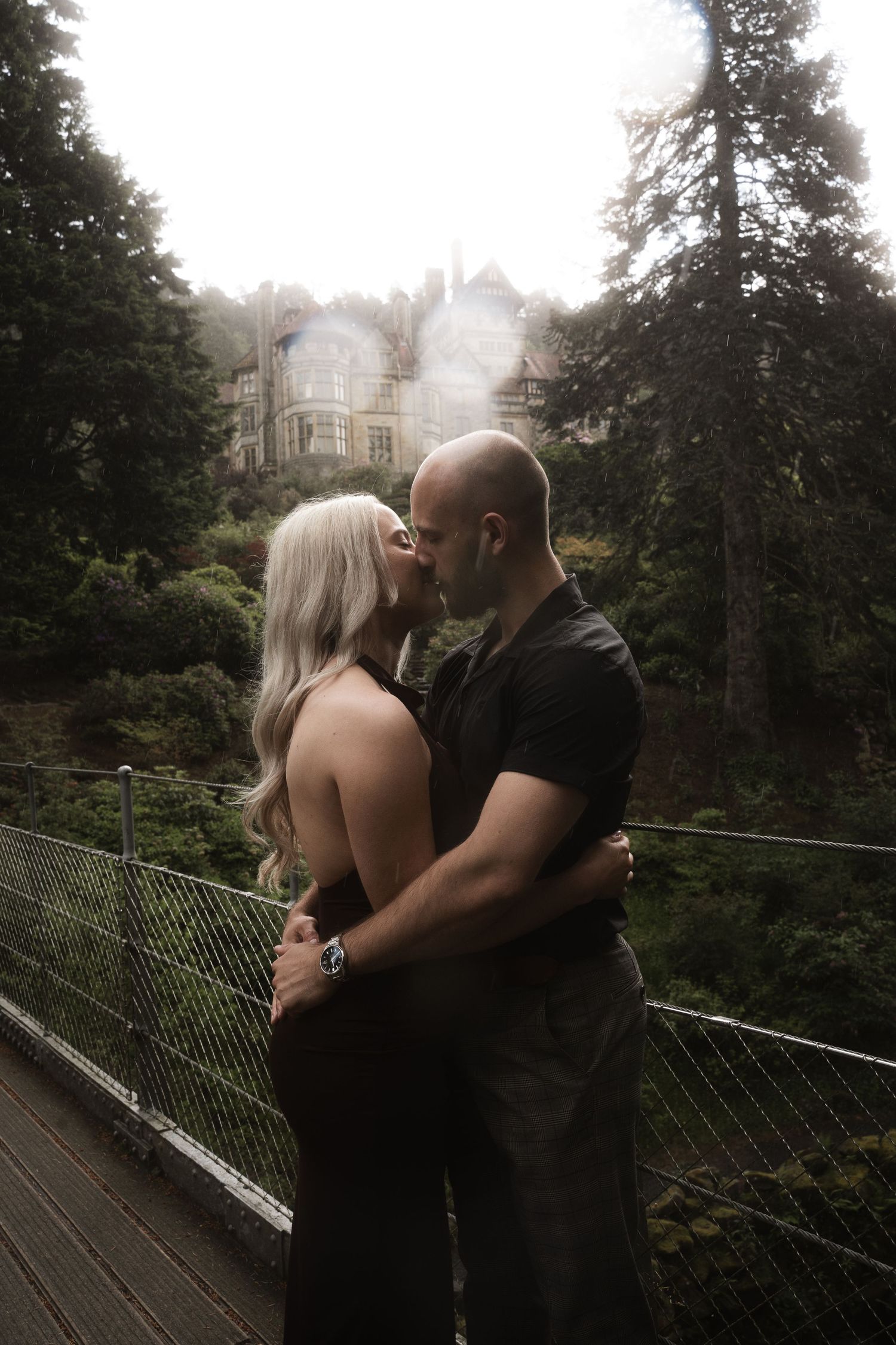 A couple shares a romantic moment while embracing on a misty wooden bridge surrounded by forest trees.