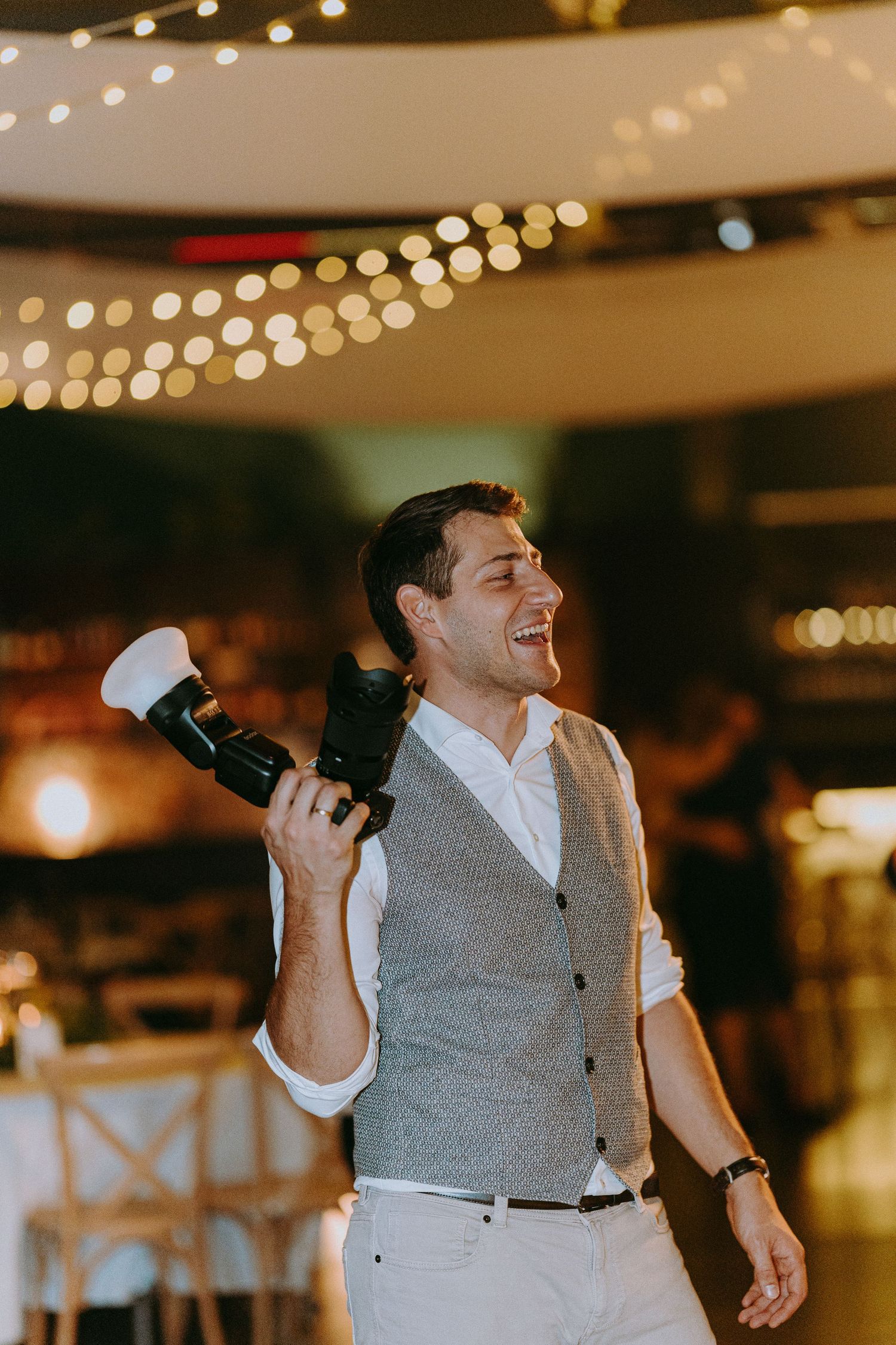 Wedding photographer in grey vest laughs while holding camera at reception with string lights.