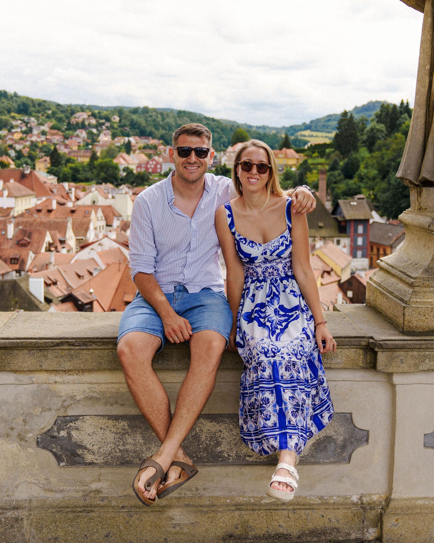 Tourist couple relaxing on stone ledge above picturesque European town.