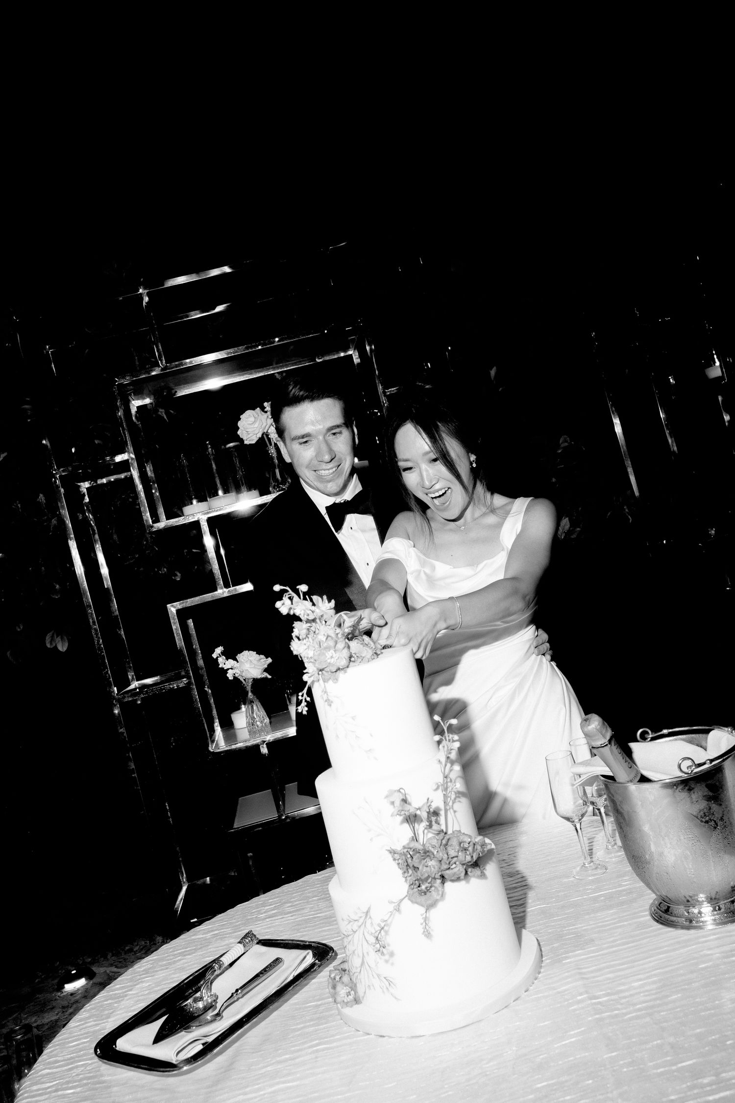 A couple shares a joyful moment while cutting their elegant white wedding cake at Pyrgos Petreza, Athens, Greece.