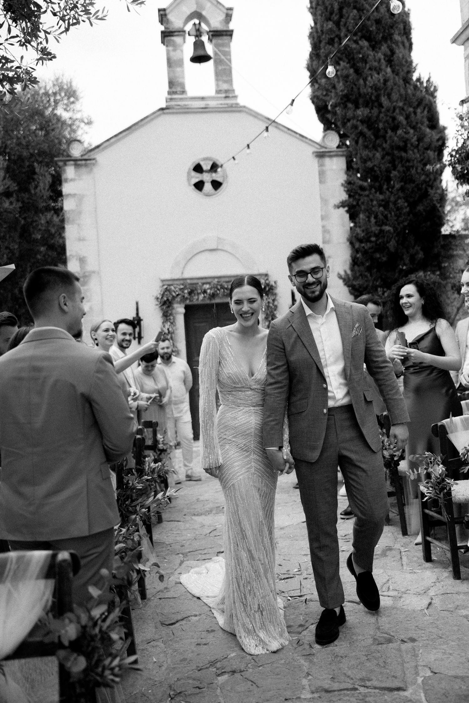 Newlyweds walk hand in hand down chapel steps while wedding guests capture the joyful moment. Agreco Farms, Rethymno.