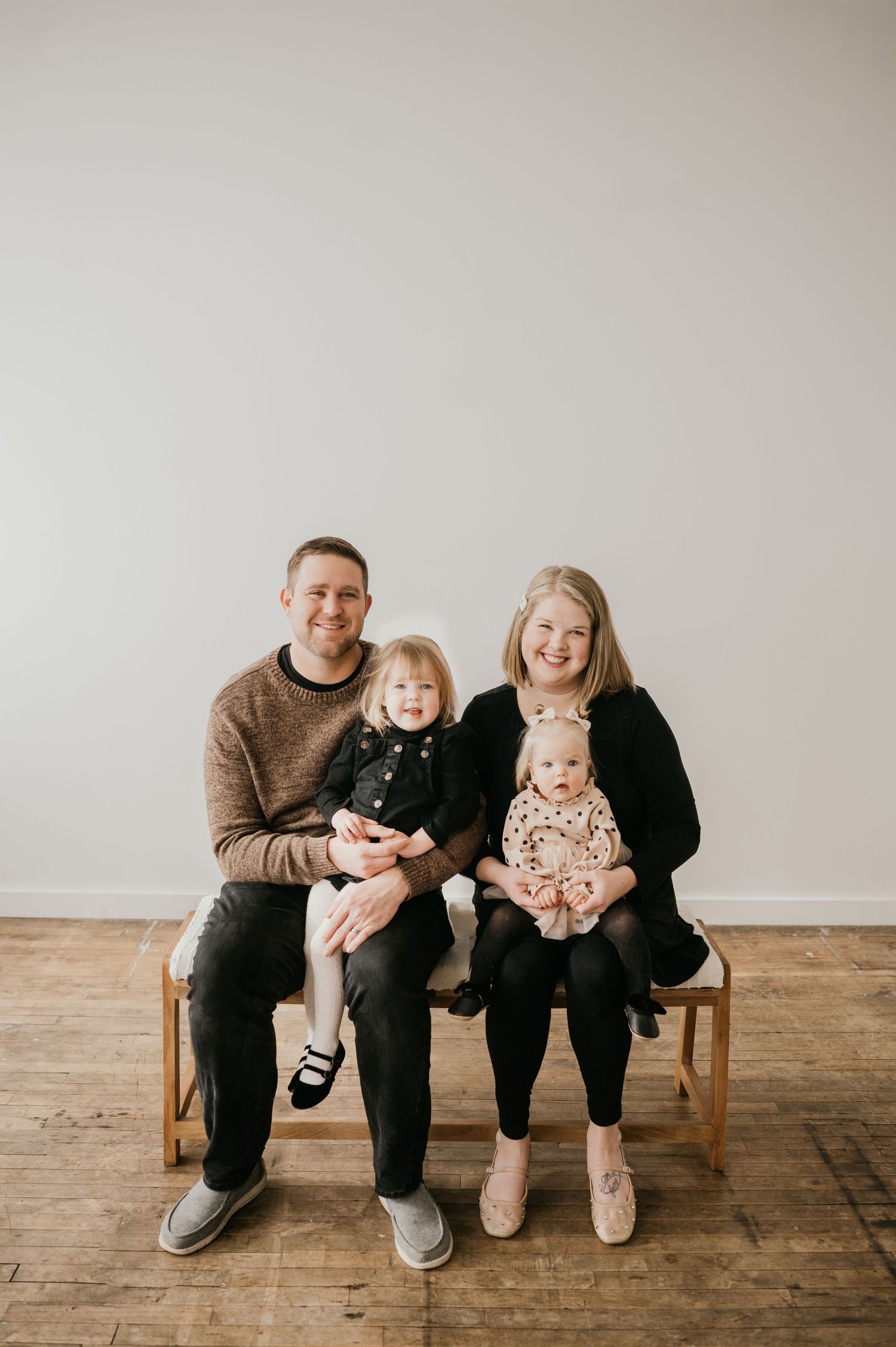 A family sitting together on a wooden bench against a white wall in a minimalist studio setting.