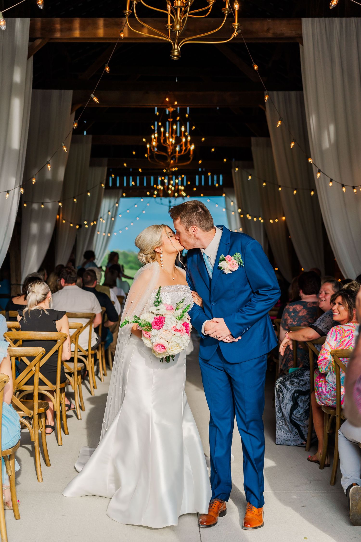 Bride and Groom kissing at the end of the aisle walking out of a barn with fairy lights behind them