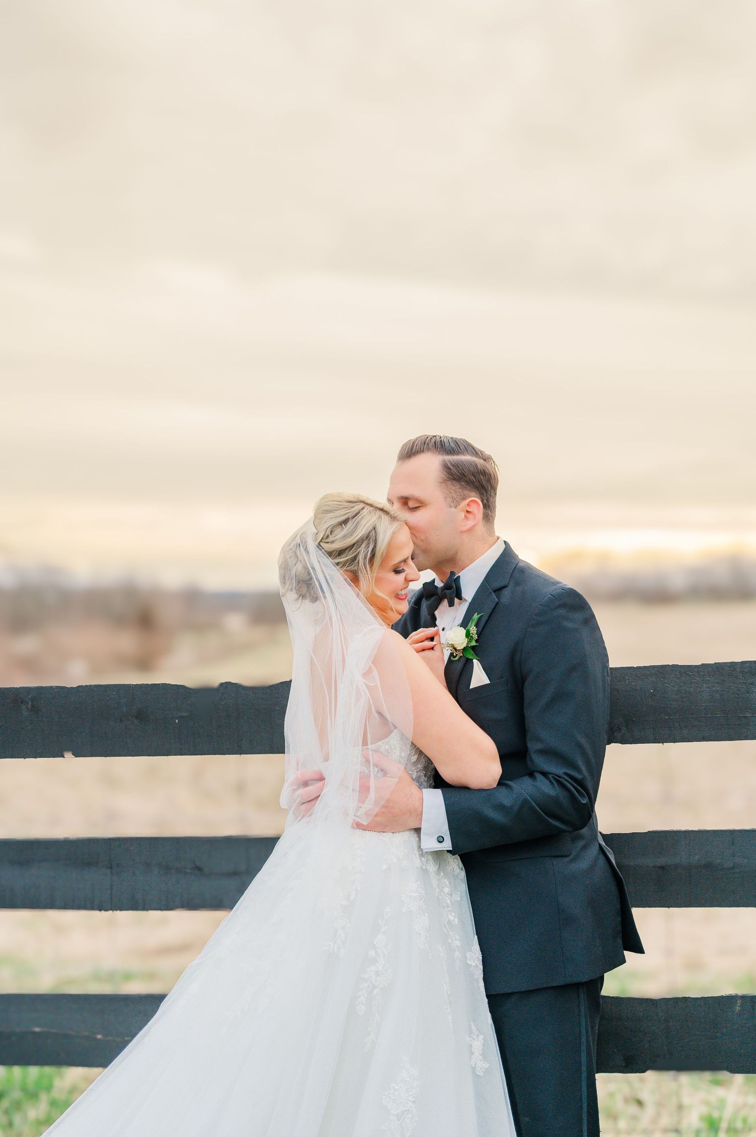 Groom kissing Brides temple in front of a fence with sunset in the background