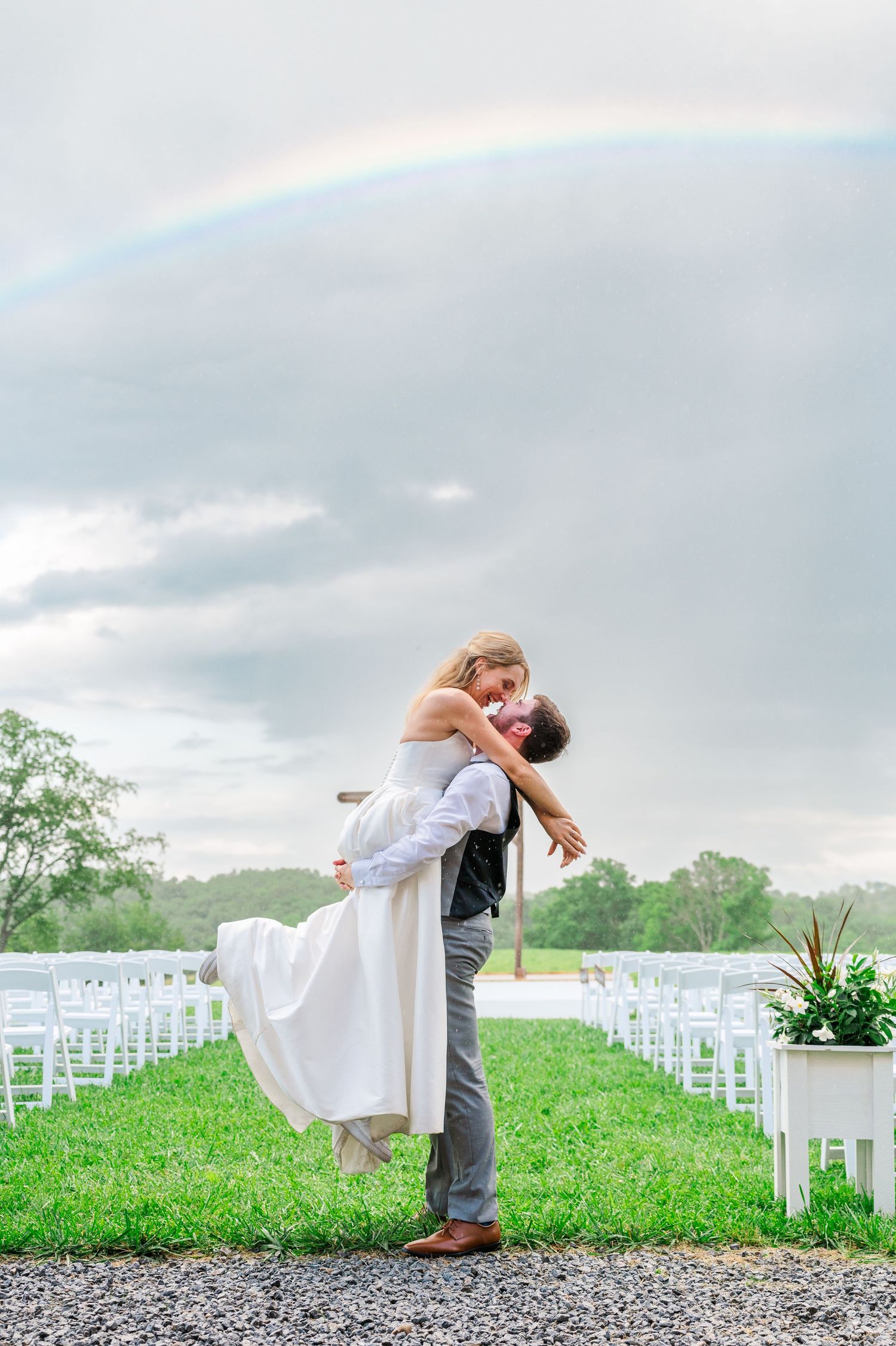 Groom picking up bride and kissing her in front of storm clouds and a rainbow