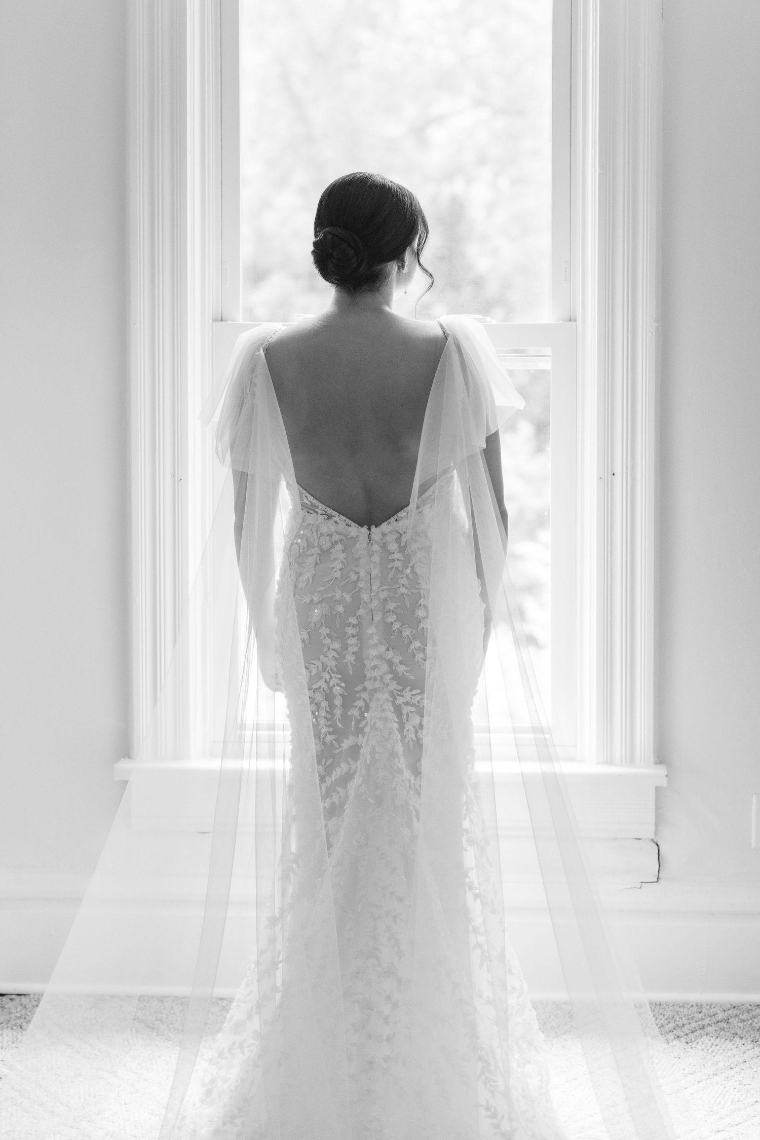 Elegant white lace wedding dress with open back shown from behind against bright window.