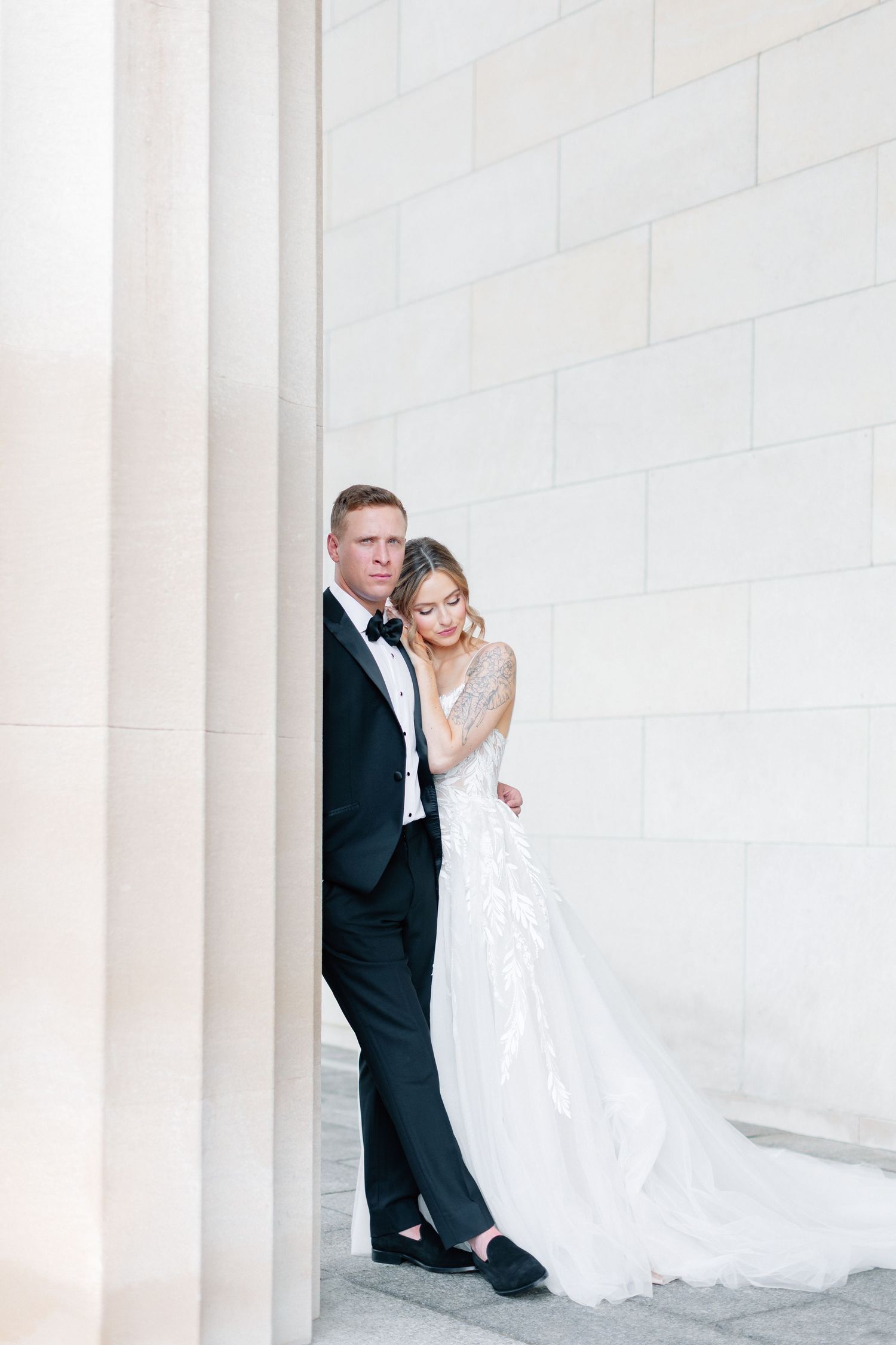 Elegant bride and groom pose intimately against white stone wall in formal wedding attire.