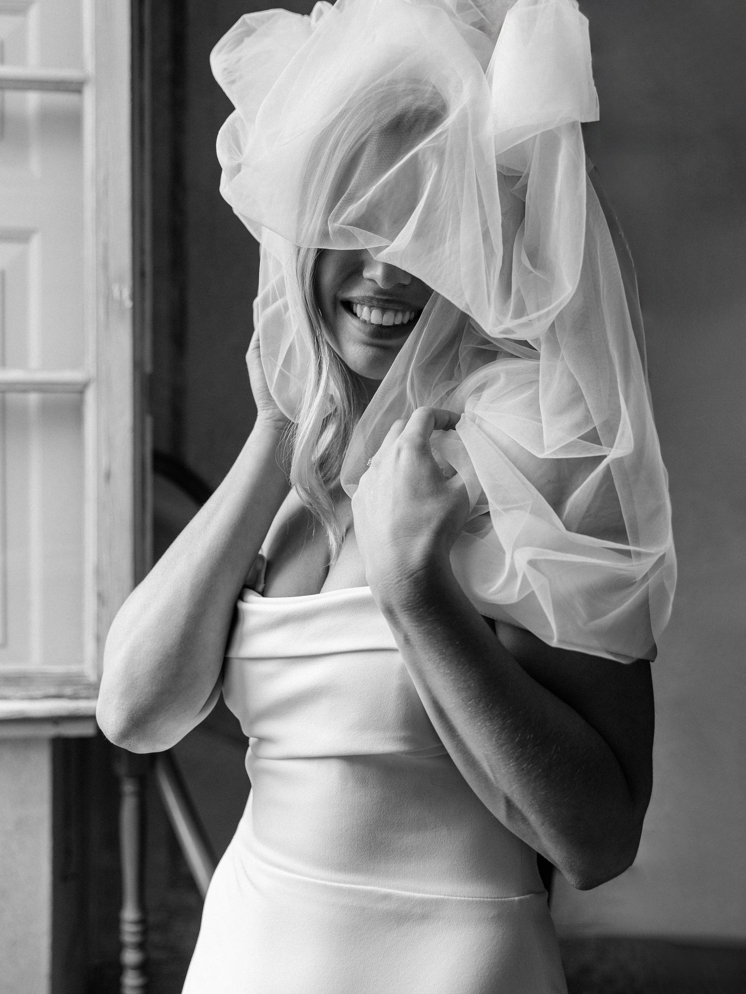 A joyful bride wearing a white wedding dress playfully covers her face with a flowing veil in black and white photography.