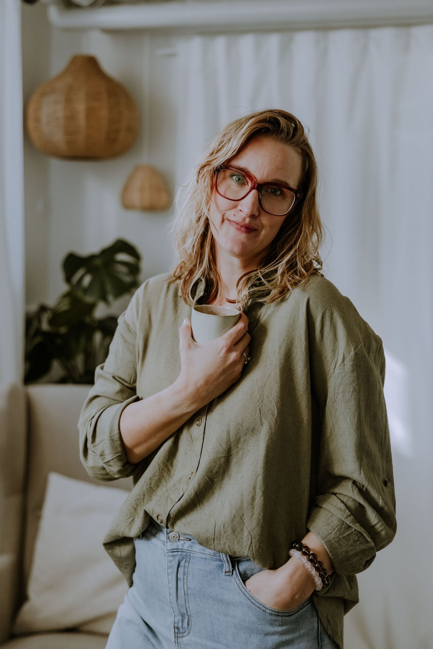 A stylish woman in glasses and olive jacket poses casually against a white wall with pendant lights and plants.