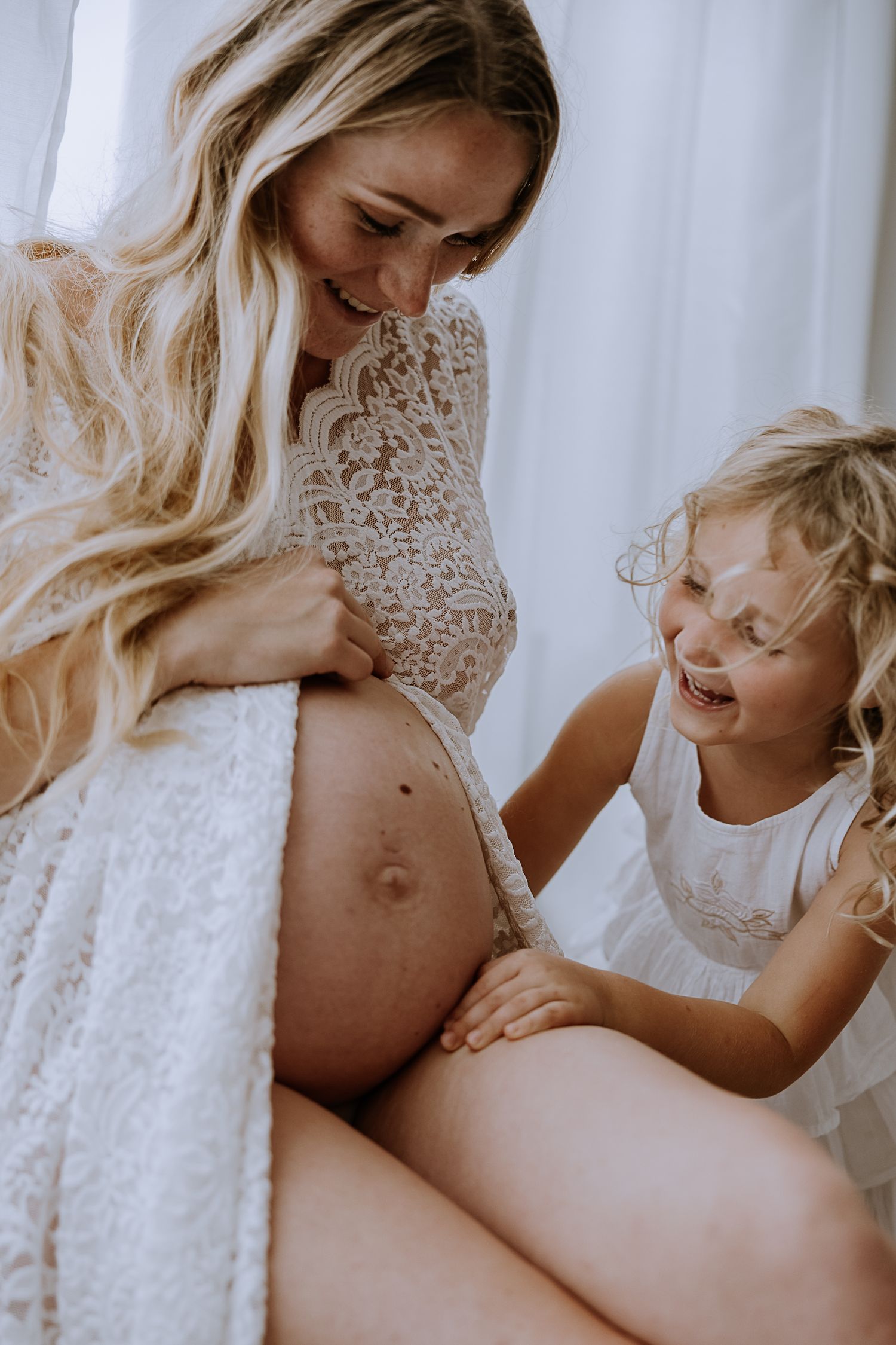 Tender moment between expecting mother in lace and a child laughing while touching her pregnant belly.