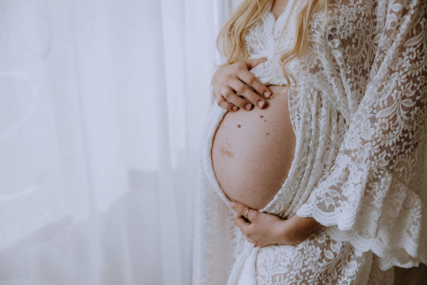 A pregnant woman's silhouette in a white lace dress gently cradles her belly against a light background.