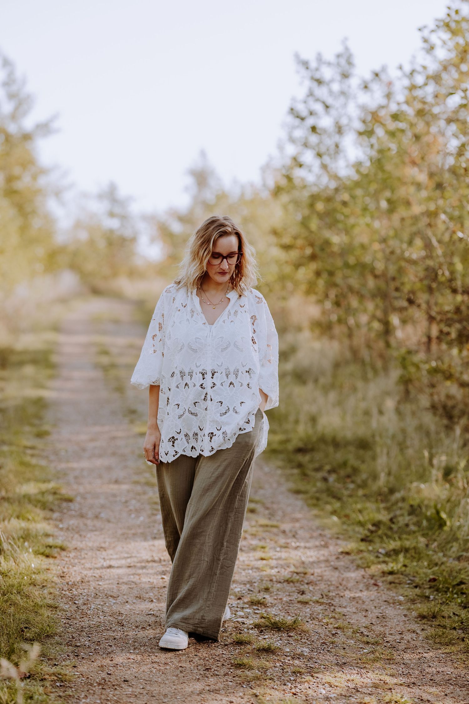 Someone in a flowing white top and wide-leg pants walks along a nature path surrounded by greenery.