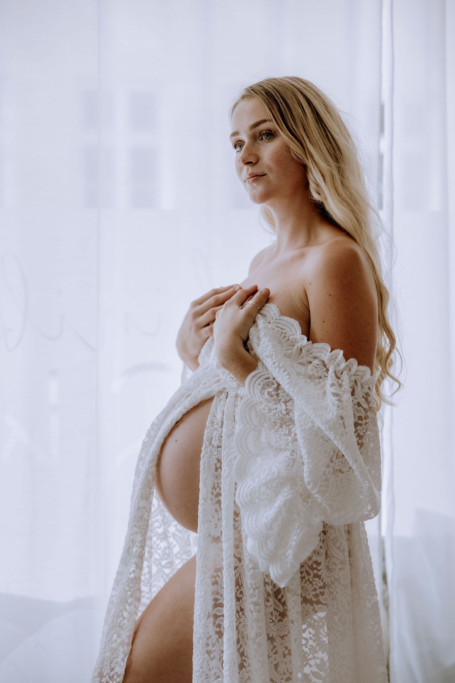 Artistic maternity photo with lace robe against white curtains in soft natural window light.
