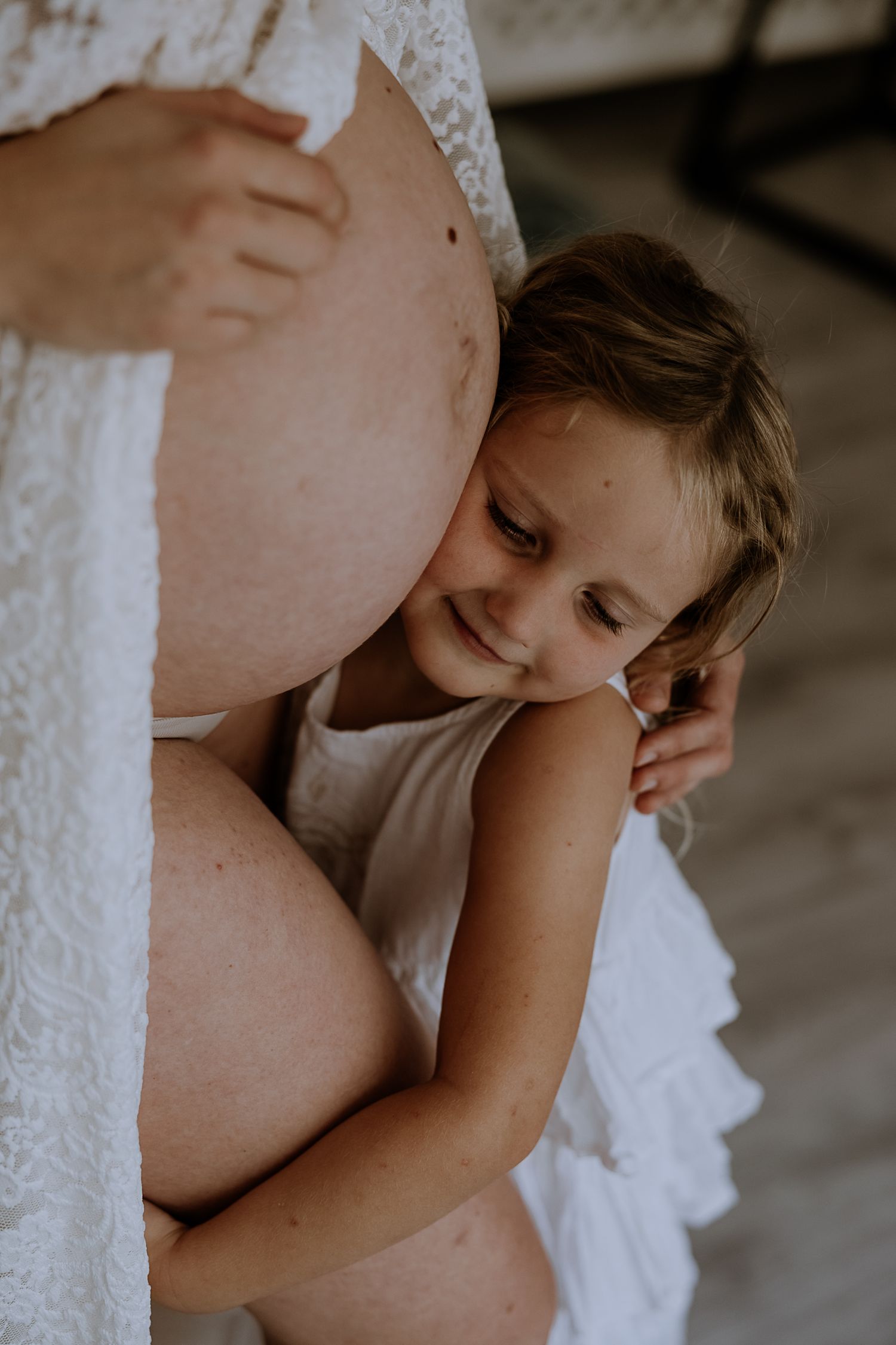 Child in white tank top rests head against pregnant belly in tender maternity moment with lace dress details.