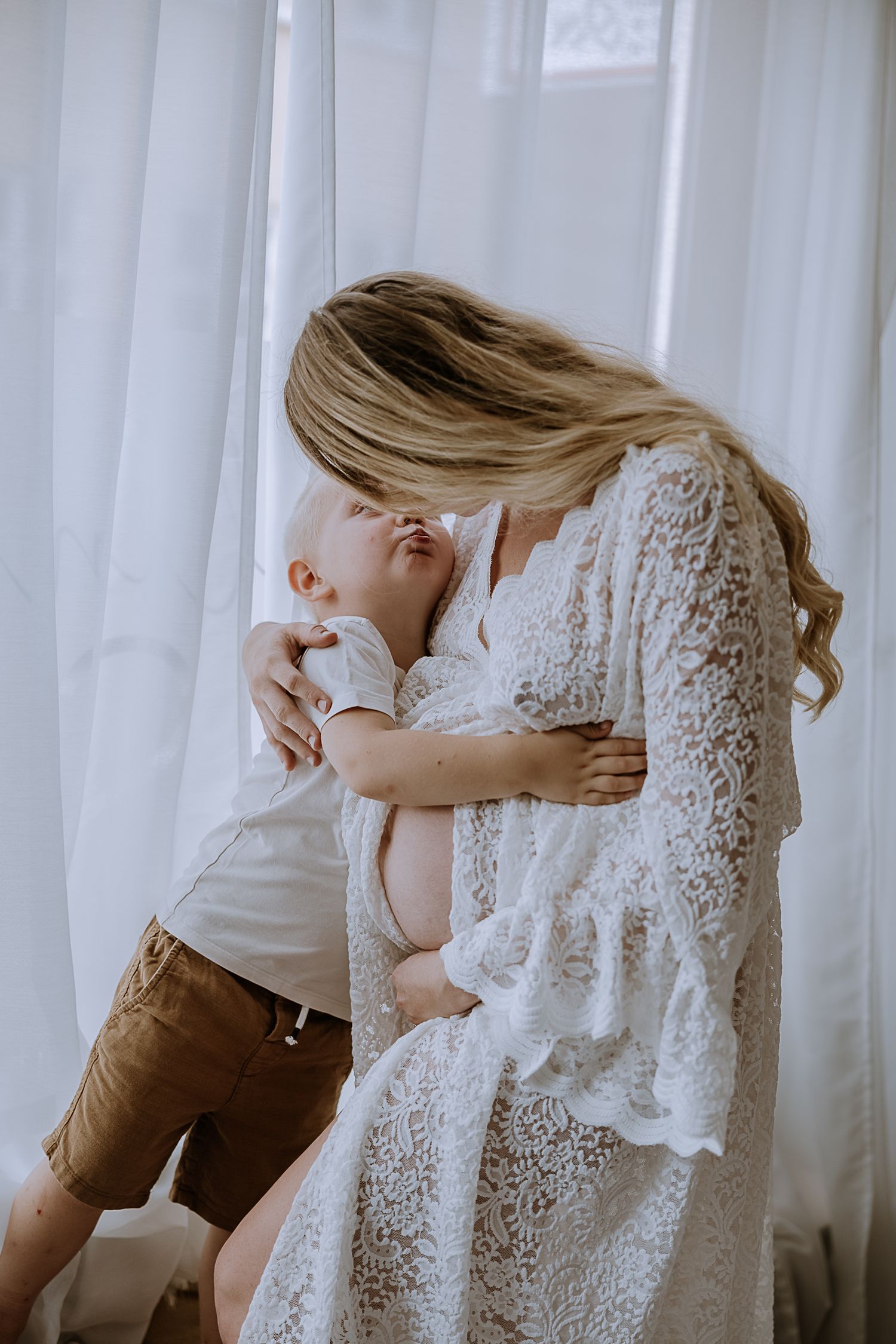 Intimate moment between mother in white lace dress and playful child in brown shorts near sheer curtains.