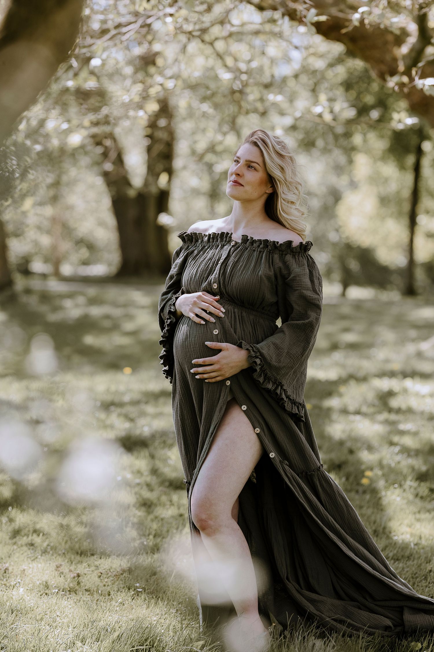 Elegant maternity portrait in a flowy black off-shoulder dress against dreamy outdoor garden setting with soft natural light.