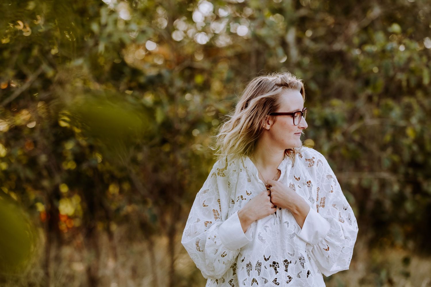 Someone wearing a delicate white lace blouse poses outdoors with golden autumn foliage in background.