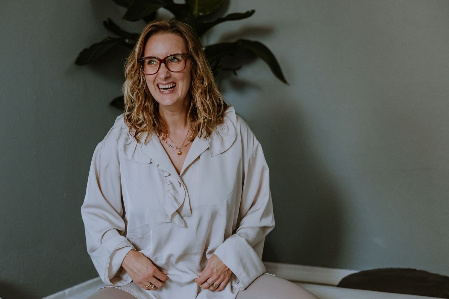 A person in white loungewear sits relaxed against a grey wall with a houseplant.